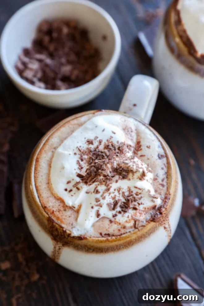 A steaming mug of dark chocolate hot chocolate, adorned with whipped cream and delicate chocolate shavings, sits invitingly on a rustic wooden table. A bowl of finely grated chocolate rests in the soft-focus background, hinting at the rich ingredients used to create this decadent treat.