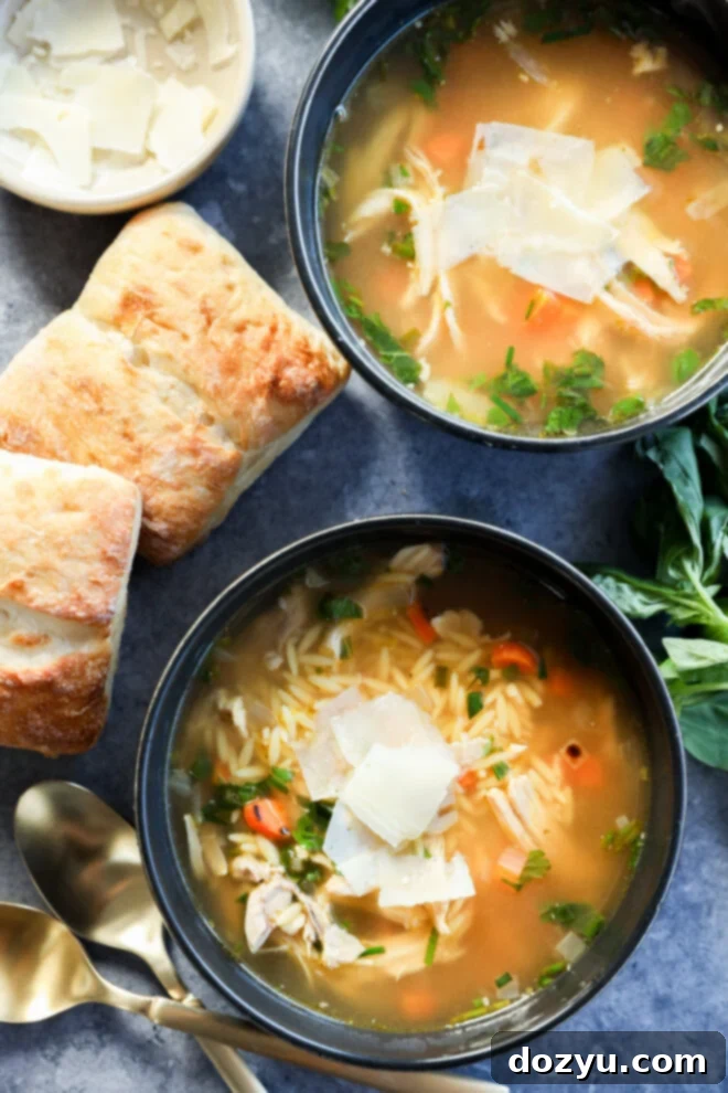 Two bowls of lemon and chicken soup with orzo, topped with shaved parmesan, are surrounded by fresh herbs, two pieces of ciabatta bread, a small bowl of parmesan, and two gold spoons on a gray surface.