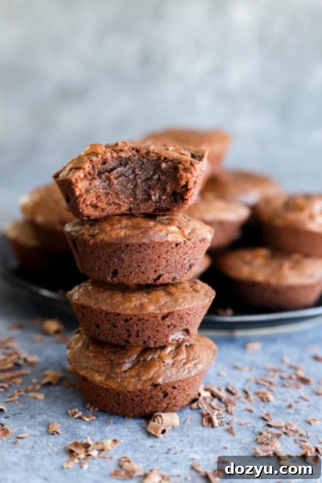 A stack of three chocolate brownies, with the top brownie missing a bite, showcases irresistible Brownie Bites. More brownies and chocolate shavings are scattered in the background on a gray surface.