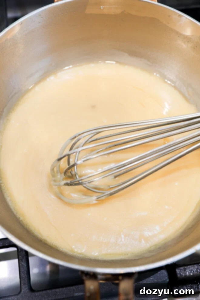 A metal whisk stirs a light tan roux in a saucepan on the stovetop, creating a smooth, slightly glossy mixture