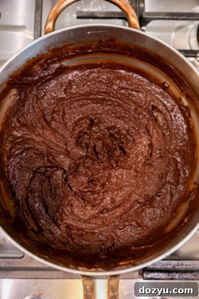 A pot on a stovetop filled with thick, dark brown chocolate mixture being stirred. The glossy texture and swirling patterns