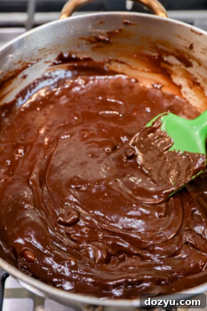 Thick, glossy chocolate brownie bites batter is being stirred in a saucepan with a green spatula on a stovetop.
