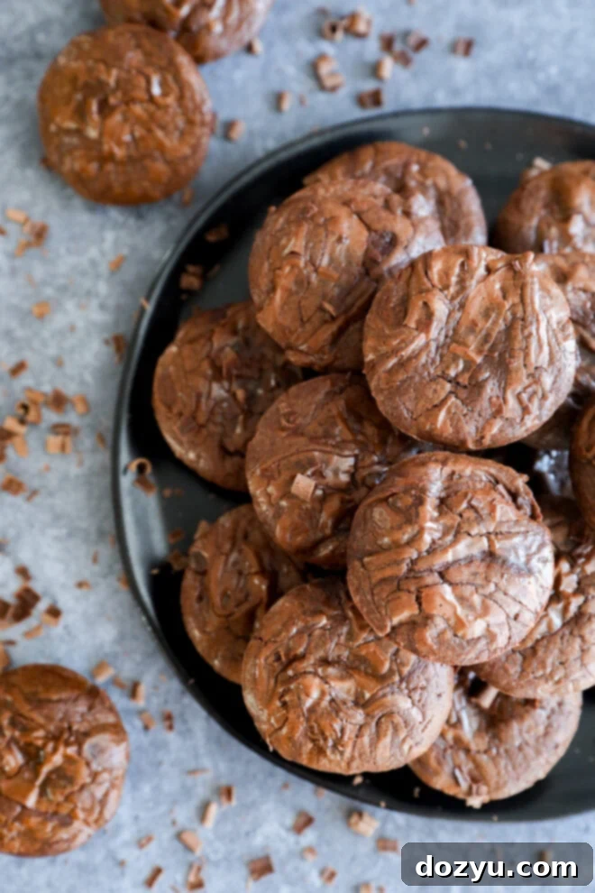 A black plate stacked with fudgy chocolate cookies and Brownie Bites, topped with chocolate shavings. More cookies and shavings are scattered on the gray surface around the plate.