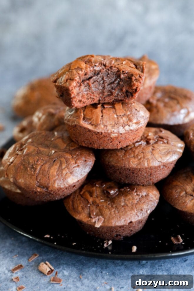 A stack of rich, fudgy chocolate brownies is displayed on a black plate. The top brownie bite has a chunk missing, revealing its moist, gooey center. Chocolate chunks are scattered on the plate and surface nearby.