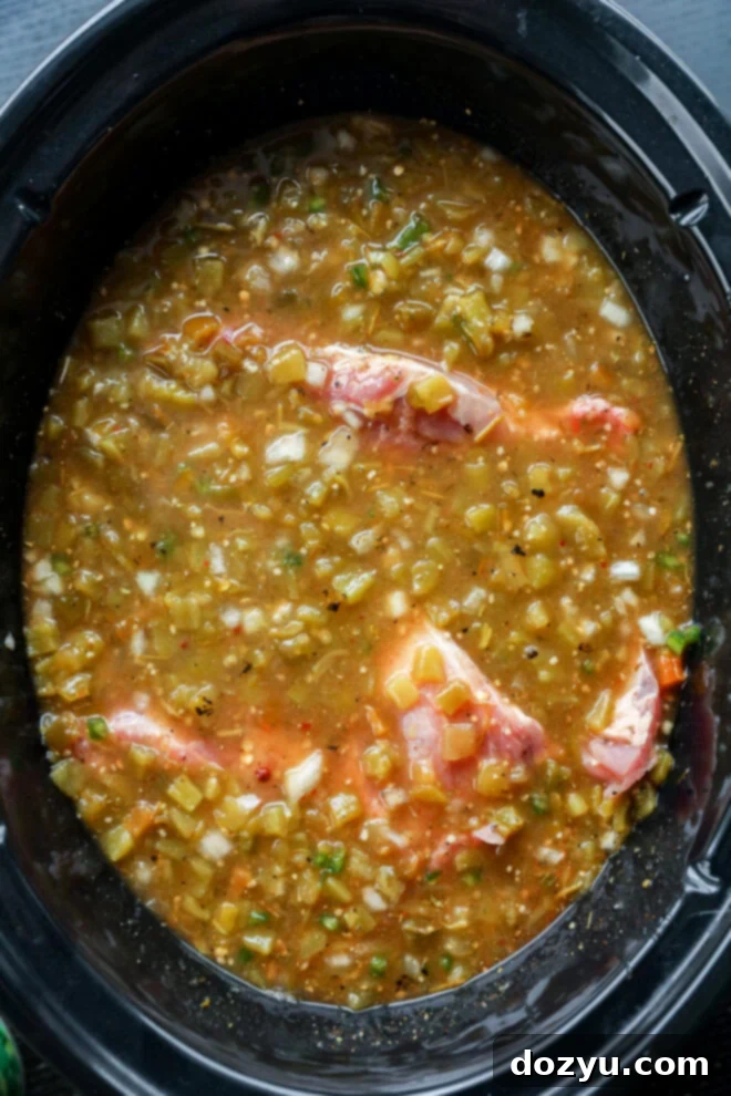 Raw pork chops are coated in a chunky pork green chile salsa with diced vegetables, all nestled in a black slow cooker and ready to be cooked.