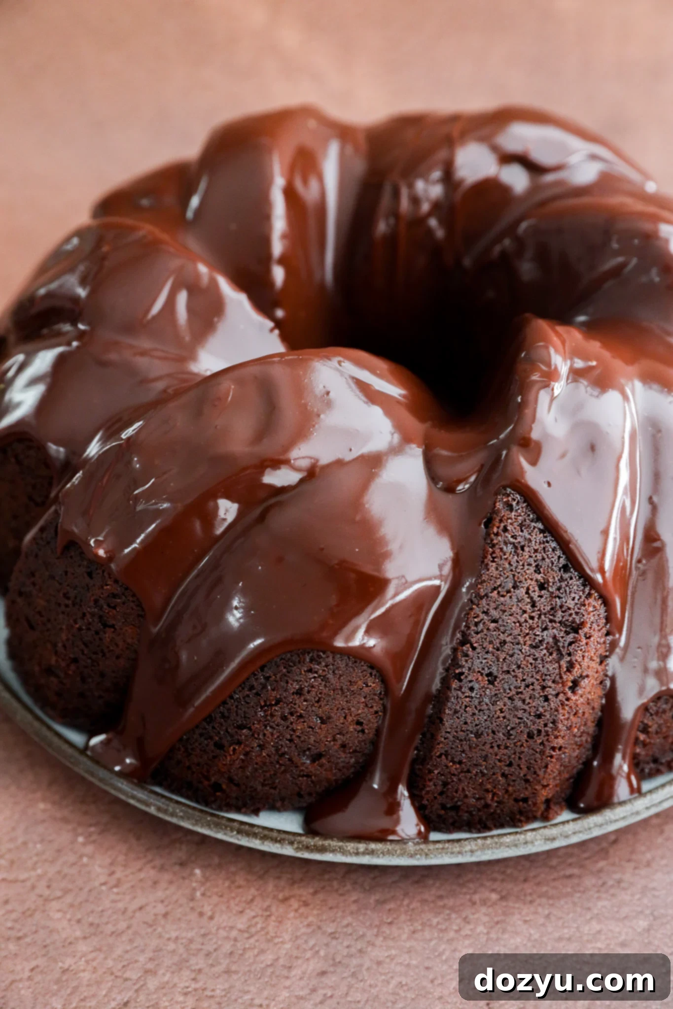 A rich brownie cake in bundt form, covered with glossy chocolate glaze, sits on a round metal plate against a brown background.