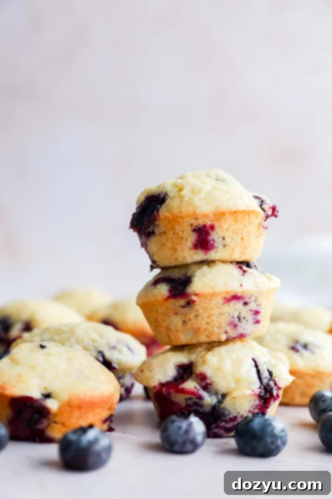 Tiny Blueberry Bites 2 A stack of three Mini Blueberry Muffins is in focus, with more muffins and scattered fresh blueberries around them on a light surface. The background is softly blurred.