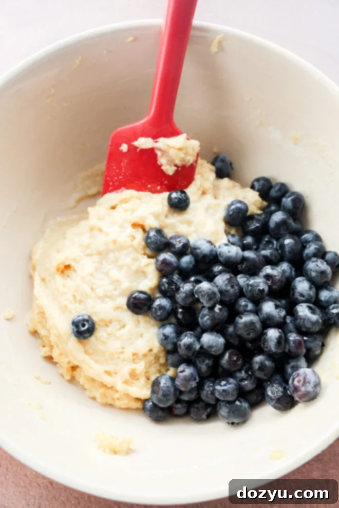 Tiny Blueberry Bites 5 A mixing bowl with mini blueberry muffins batter and fresh blueberries, being stirred with a red spatula.