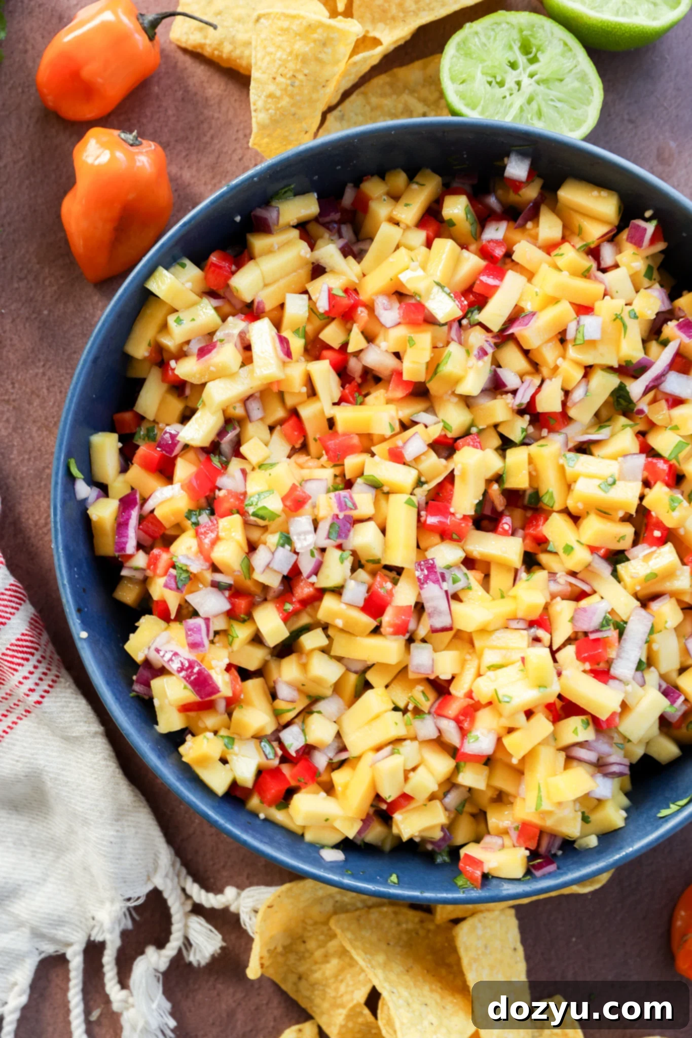 A bowl of mango salsa with diced mango, red onion, red bell pepper, and herbs, surrounded by tortilla chips, a halved lime, habanero peppers, and a white cloth on a brown surface.